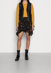 Frau in senfgelbem Pullover, schwarzem Lederweste, asymmetrischem Blumenrock und schwarzen Ankle Boots lehnt an einer weißen Wand.