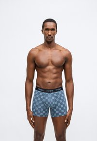 Cuecas boxer masculinas em azul com um padrão repetido do logo "HUGO", apresentando uma cintura elástica preta. Material em algodão com textura suave.