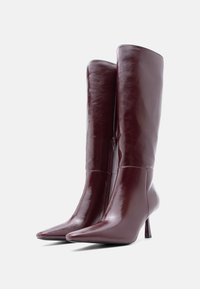 Botas de cuero burdeos hasta la rodilla con acabado brillante, puntas afiladas y un tacón alto y delgado. Presenta un zipper lateral para facilitar su uso.