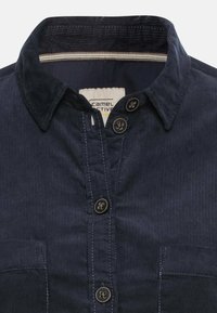 Granatowa koszula z korduroy z zapięciem na guziki, dwoma kieszeniami na piersi i kołnierzykiem z wycięciem. Cechuje się widocznymi czarnymi guzikami oraz kontrastowymi przeszyciami.