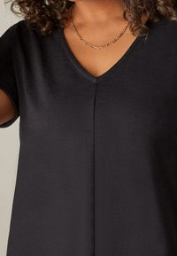 Svart kortärmad topp med V-ringning gjord av mjukt tyg, med en central söm ned längs framsidan. Elegant smal kedjesmycke synligt.