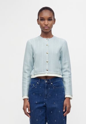 Jeune femme aux cheveux tirés en arrière portant un cardigan en tricot bleu clair boutonné et un jean bleu foncé avec de petits motifs blancs.