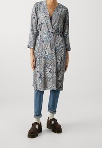 Wzorzysta, długorękawowa sukienka kopertowa z motywem paisley w kolorach niebieskim i beżowym, zestawiona z jasnoniebieskimi jeansami i brązowymi butami.