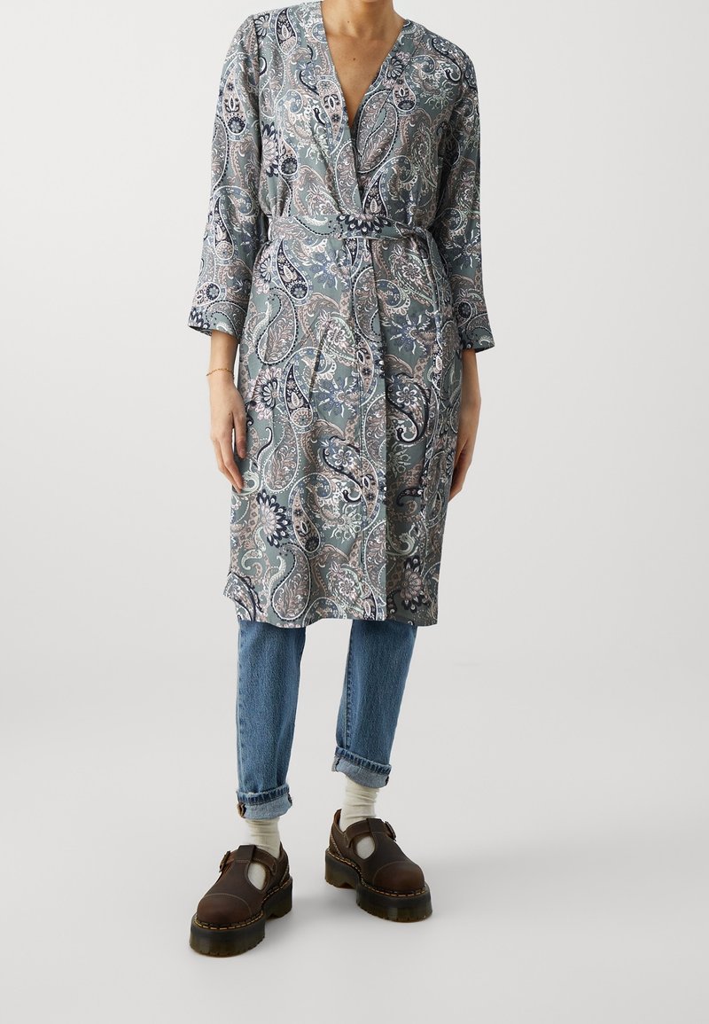 Wzorzysta, długorękawowa sukienka kopertowa z motywem paisley w kolorach niebieskim i beżowym, zestawiona z jasnoniebieskimi jeansami i brązowymi butami.