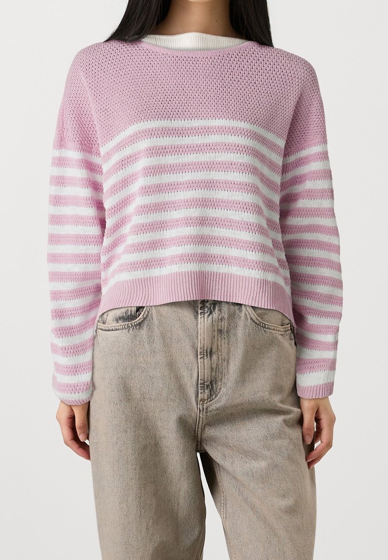 Pull en tricot rayé rose et blanc avec un col rond et un ourlet côtelé. Associé à un pantalon en denim de couleur claire.