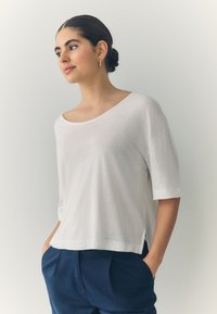 White short-sleeve top made of soft fabric with a wide neckline and side slits, paired with dark blue trousers. Simple, modern design.