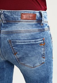 Jeans de mezclilla azul descoloridos con un ligero estiramiento, con costuras marrones, un parche de cuero etiquetado "LTB" y detalles en el bolsillo trasero.