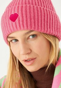 Roze geribde gebreide beanie met een klein geborduurd hart. Benadrukte textuur en een comfortabele pasvorm. Achtergrond met een kleurrijke sjaal.