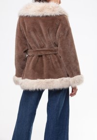 Cappotto in pelliccia sintetica marrone con un ampio collo e polsini in pelliccia chiara, vita cintrata e fondo svasato. Indossato sopra jeans a gamba larga blu scuro.