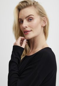 Črn dolg rokav majica z okroglo ovratnico in rebrastimi manšetami, ki jo nosi oseba z dolgimi, valovitimi blond lasmi. Gladka tekstura in prilegajoč kroj.