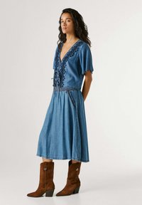 Vrouw draagt een blauwe spijkerjurk met geborduurde details en bruine suède laarzen tot halverwege de kuit, staand tegen een effen achtergrond.