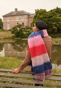 Farebná vlnená šatka s výraznými pruhmi v červenej, ružovej a modrej farbe. Mäkká textúra s strapcovými koncami a detailom kruhového loga.