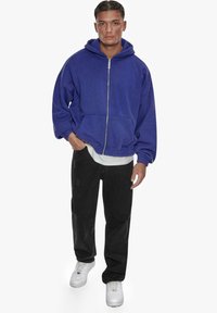 Haut à capuche zippé bleu avec cordon de serrage, coupe oversize, associé à un pantalon noir et des baskets blanches. Tissu texturé, design décontracté.