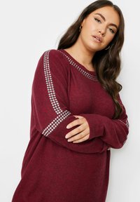 Burgundy sweatshirt med mjuk textur, prydd med silverfärgade nitar längs axlarna och långa ärmar. Avslappnad passform med rund halsringning.