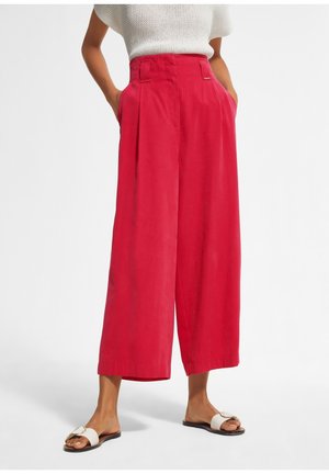 Femme portant un pantalon rouge taille haute à jambes larges avec poches latérales, associé à un top en maille blanche et des sandales plates blanches.