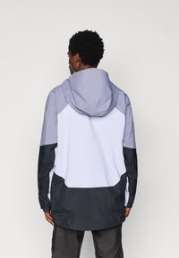 Άνδρας με καπιτονέ μπουφάν με κουκούλα σε λεβάντα, άσπρο και μαύρο, στέκεται με την πλάτη στραμμένη προς την κάμερα μπροστά σε απλό φόντο.