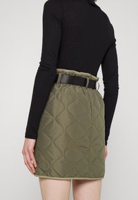 Jupe en olive matelassée avec une taille ceinturée noire, arborant un motif ondulé. Le tissu semble lisse et léger.
