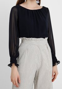 Blusa nera trasparente con scollatura plissettata e maniche lunghe a balze abbinata a pantaloni a vita alta con righe.