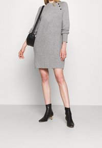 Abito grigio lavorato a maglia con scollo alto, caratterizzato da tre bottoni neri sulla spalla. Indossato con stivali neri alla caviglia.