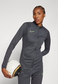 Gray sporty zip-up jacket with long sleeves and Nike logo highlighted in neon green. Model holds a textured white and black soccer ball.