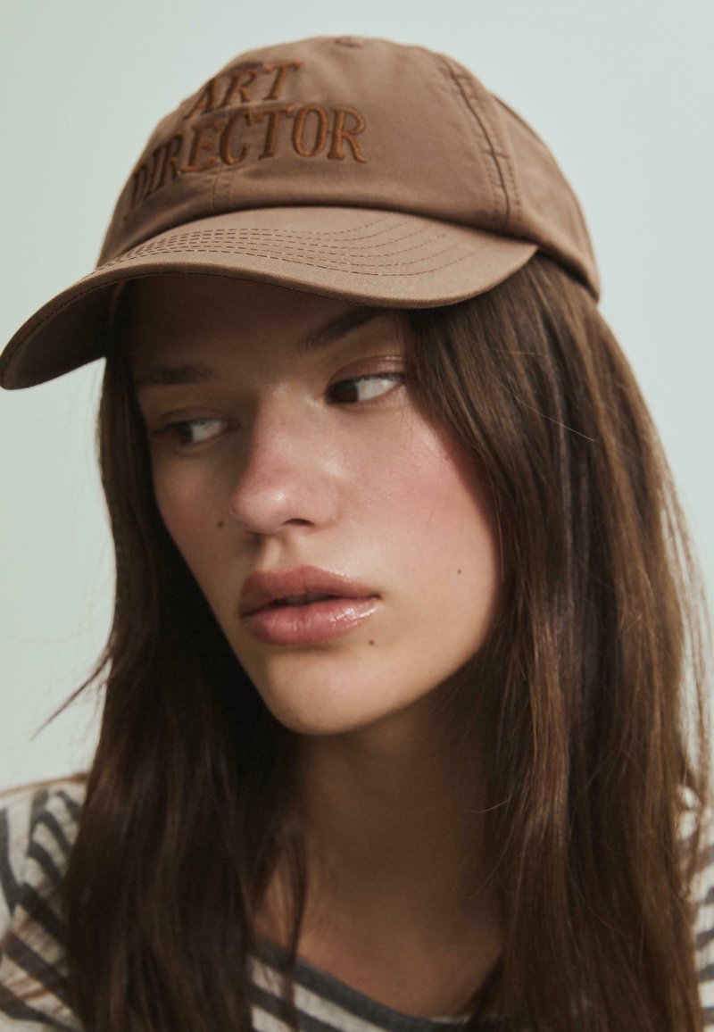 Brun baseball cap med "ART DIRECTOR" broderet i matchende tråd; har en buet skygge og tekstureret stof. Stribet skjorte synlig nedenunder.