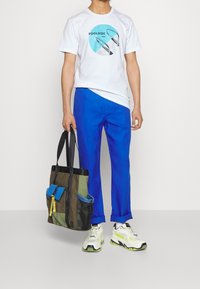 Bílý tričko s modrofialovou grafikou, modré kalhoty a vícebarevná taška s kapsami; bílá a zelená sneakers.