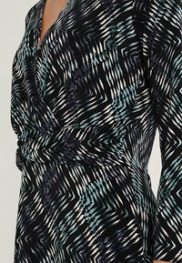 Långärmad klänning i svart tyg med ett geometriskt mönster i teal, vitt och blått. Designen har en inlindad midjedetalj.
