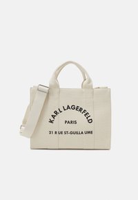 Beige canvas toteväska med dubbla handtag, justerbar rem och svarta tryckta varumärkesdetaljer. Har en strukturerad design och dragkedja.