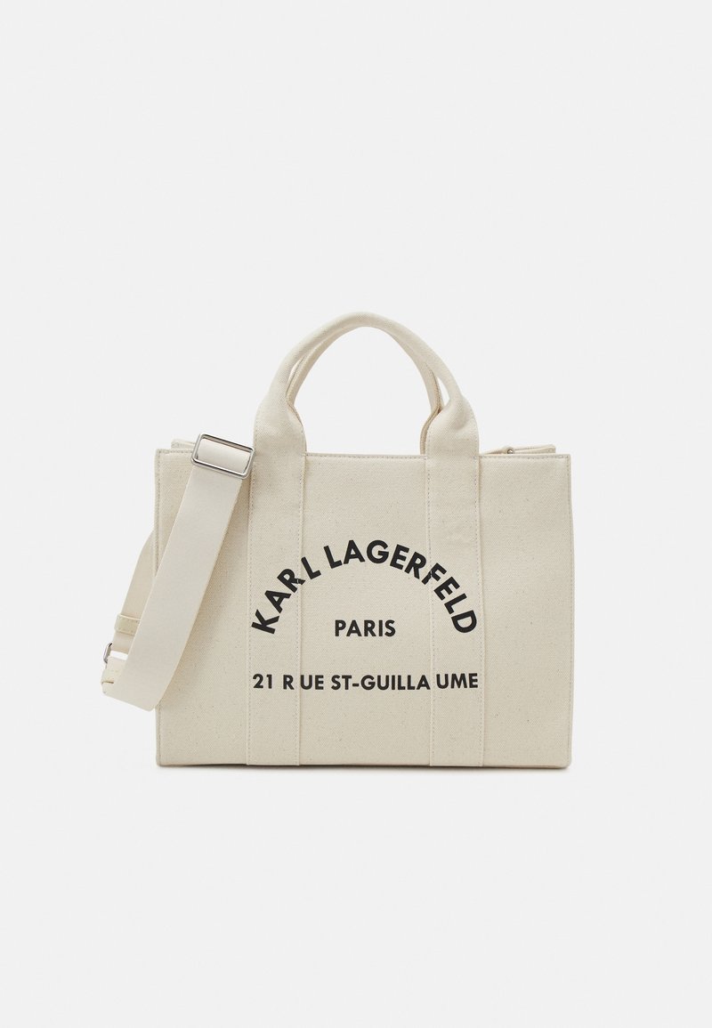 Beige canvas toteväska med dubbla handtag, justerbar rem och svarta tryckta varumärkesdetaljer. Har en strukturerad design och dragkedja.