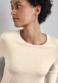 Beige svetr s kulatým výstřihem, měkkou strukturou a přiléhavým střihem. Modelka nosí stříbrné kruhové náušnice, což ukazuje na casual vzhled.
