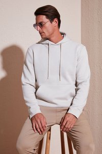Felpa con cappuccio grigio chiaro con tasca frontale, cordini e polsini elasticizzati, abbinata a pantaloni beige. Modello seduto su uno sgabello di legno.