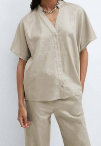 Chemise beige clair à manches courtes, à boutons, avec un tissu texturé, un col en V et une légère brillance. Associée à un pantalon assorti. Collier en chaîne argentée.