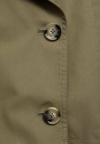 Gros plan de deux boutons ronds marbrés beige et noir fixés sur un tissu vert olive avec des coutures visibles le long du bord.