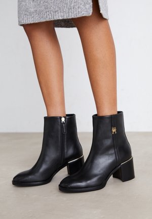 Jambes portant des bottines en cuir noir à talons carrés, avec des zippers sur le côté et un détail en logo doré, associées à une jupe en tricot grise.