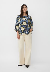 Blusa de diseño floral, de corte holgado, en azul, amarillo y crema, combinada con pantalones de pierna ancha en beige y zapatos de punta negra.