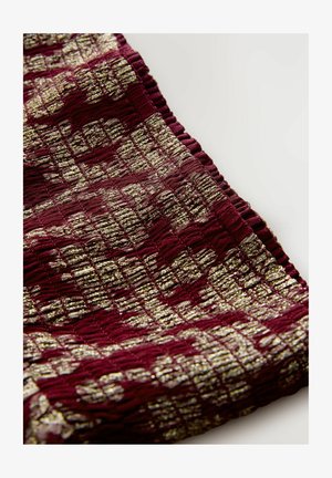 Närbild av texturerad tyg med en mörkröd botten och ett metalliskt guldrektangulärt mönster, med en volangkant på en ljus bakgrund.