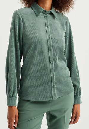 Kobieta ubrana w zieloną sztruksową koszulę z długim rękawem zapinaną na guziki oraz zielone spodnie, stojąca z luźno opuszczonymi rękami.