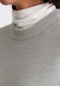 Maglione grigio chiaro testurizzato con scollo rotondo, indossato sopra un dolcevita bianco. Il tessuto appare morbido e traspirante, caratterizzato da un motivo a maglia a nido d'ape.