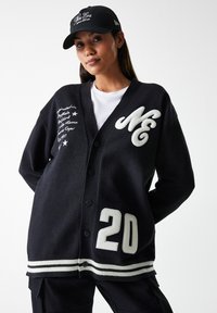 Cardigan en tricot noir avec des accents blancs, présentant un grand logo "NE", du texte brodé et un "20" bien en vue sur le bas devant.