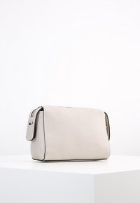 Borsa a tracolla rettangolare in pelle beige con pattina e rifiniture nere, appoggiata su una superficie bianca con sfondo bianco uniforme.
