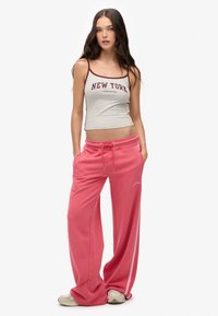Donna con lunghe chiome scure che indossa una canotta grigia di New York, pantaloni morbidi rosa a gamba larga e sneakers chiare, in piedi con le mani in tasca.