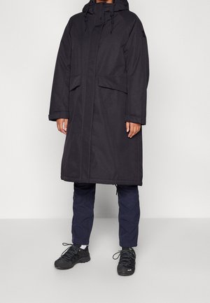 Personne portant un long manteau d'hiver noir à capuche avec des poches à rabat, un pantalon bleu marine, des chaussettes blanches et des chaussures noires lacées, debout sur un fond blanc.