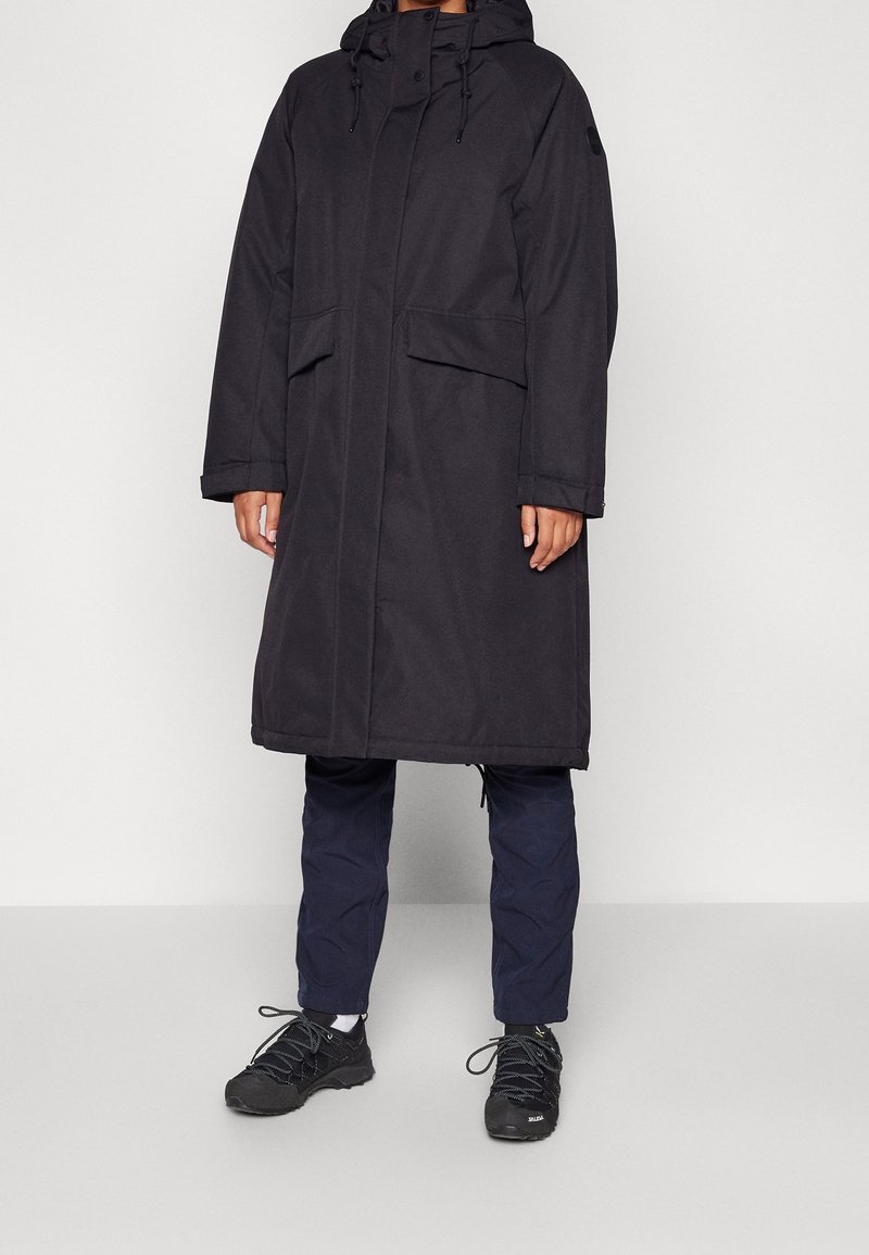 Personne portant un long manteau d'hiver noir à capuche avec des poches à rabat, un pantalon bleu marine, des chaussettes blanches et des chaussures noires lacées, debout sur un fond blanc.