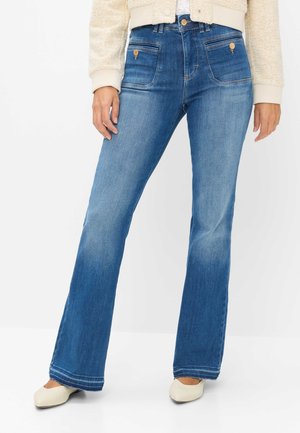 Kvinde iført blå højttaljede flare jeans, beige tekstureret jakke og hvide flade sko står mod en ensfarvet baggrund.
