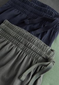 Zwei Paare von elastischen Bundhosen in dunkelmarine und olivgrün, mit Raffung und Kordelzügen, auf einer strukturierten Oberfläche.