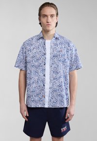 Korte mouwen overhemd in het wit met een blauw bloemenpatroon, voorzien van oranje accenten en een zak. Gecombineerd met marineblauwe shorts.