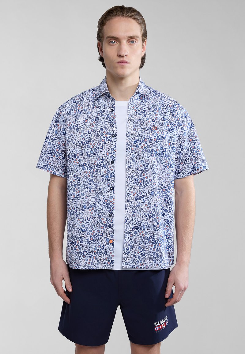 Korte mouwen overhemd in het wit met een blauw bloemenpatroon, voorzien van oranje accenten en een zak. Gecombineerd met marineblauwe shorts.