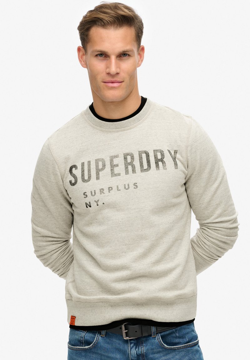 Lichtgrijs sweatshirt van stof met een soepele textuur. Het heeft een geribde hals, lange mouwen en een grafische print aan de voorkant met de tekst "SUPERDRY SURPLUS NY."
