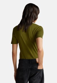 Person mit langen, dunklen Haaren, die ein olivgrünes, kurzärmliges Hemd tragen, das in hoch taillierte schwarze Jeans mit einem Lederetikett gesteckt ist.