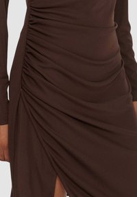 Vestito marrone con tessuto arricciato, maniche lunghe e spacco laterale per un dettaglio in più. Texture liscia con una leggera lucentezza.